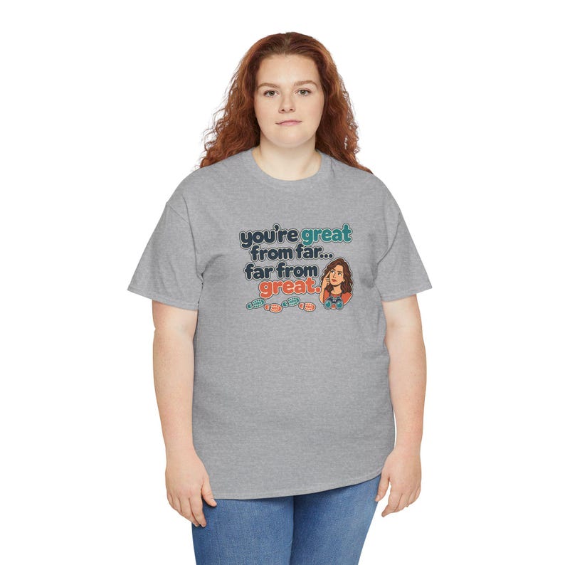 May include: A heather gray short-sleeve t-shirt featuring the text "you're great from far... far from great." with a cartoon illustration of a person looking through binoculars and small colorful footprint icons below the text.