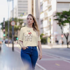 May include: A cream-colored short-sleeve t-shirt featuring a graphic illustration of a white poodle dog with a red bow on its head and the word Poodle printed above the dog in a simple black font, paired with blue denim jeans.