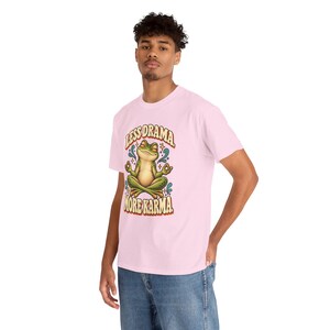 May include: A light pink t-shirt featuring a graphic of a green frog sitting in a lotus position with its eyes closed. The text above the frog reads "LESS DRAMA" and the text below reads "MORE KARMA" in a stylized, retro-inspired font.