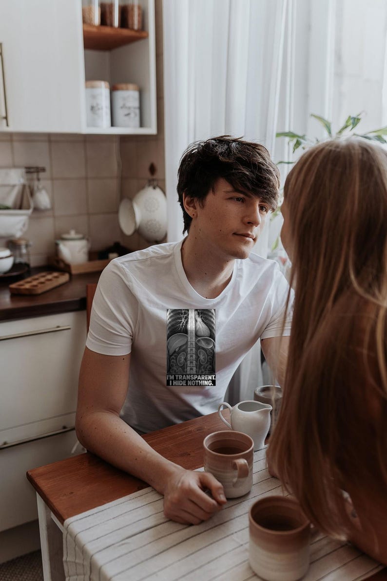 Puede incluir: Una camiseta blanca de manga corta con un gr&aacute;fico rectangular de una radiograf&iacute;a del torso humano. El gr&aacute;fico incluye el texto I'M TRANSPARENT. I HIDE NOTHING. La camiseta tiene cuello redondo y una silueta ajustada.
