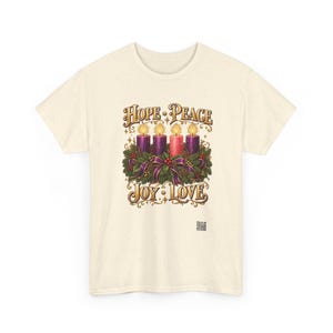 May include: A cream-colored short-sleeve t-shirt featuring a graphic of an Advent wreath with four lit candles, three purple and one pink, surrounded by greenery and ribbons. The text reads HOPE, PEACE, JOY, and LOVE in gold lettering.