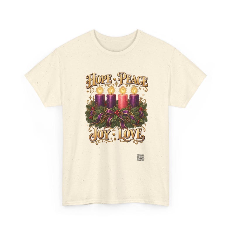 May include: A cream-colored short-sleeve t-shirt featuring a graphic of an Advent wreath with four lit candles, three purple and one pink, surrounded by greenery and ribbons. The text reads HOPE, PEACE, JOY, and LOVE in gold lettering.