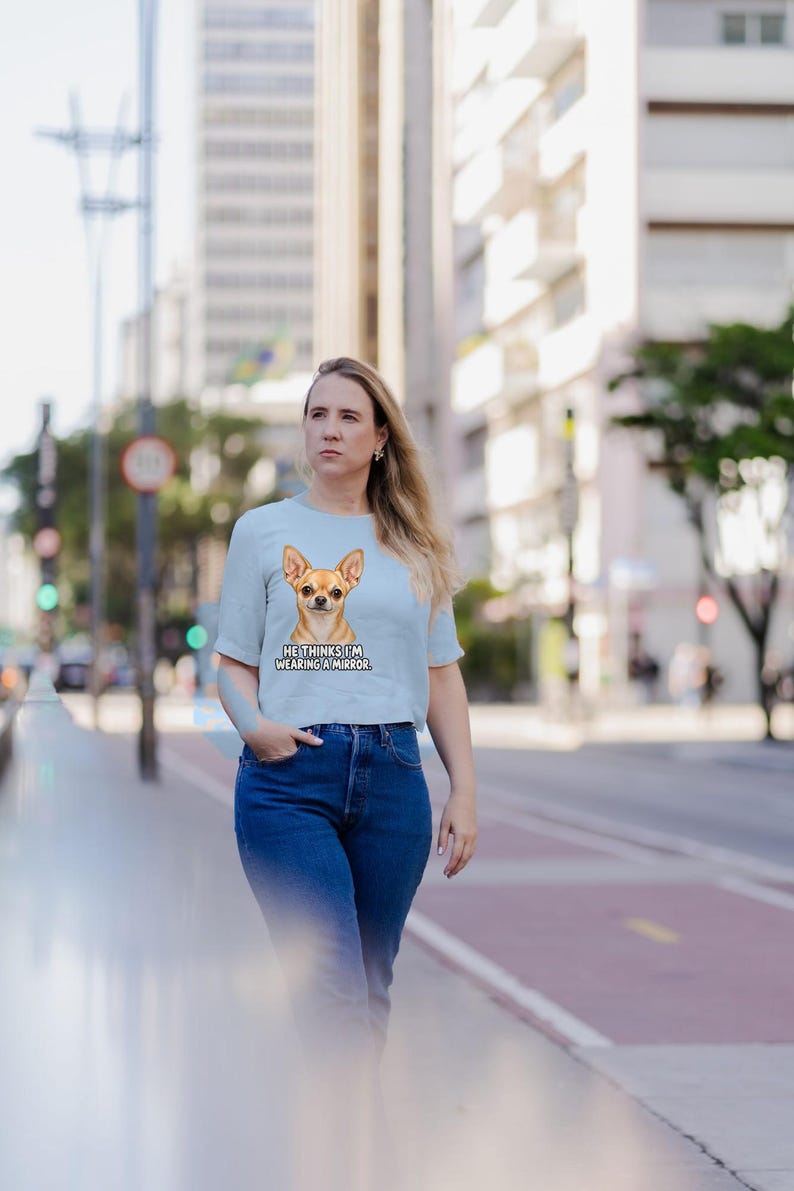 May include: A light blue short-sleeved t-shirt featuring a graphic of a tan chihuahua dog. Below the dog, the text reads, "HE THINKS I'M WEARING A MIRROR." The shirt has a crew neckline and a straight hem, worn with dark blue denim jeans.