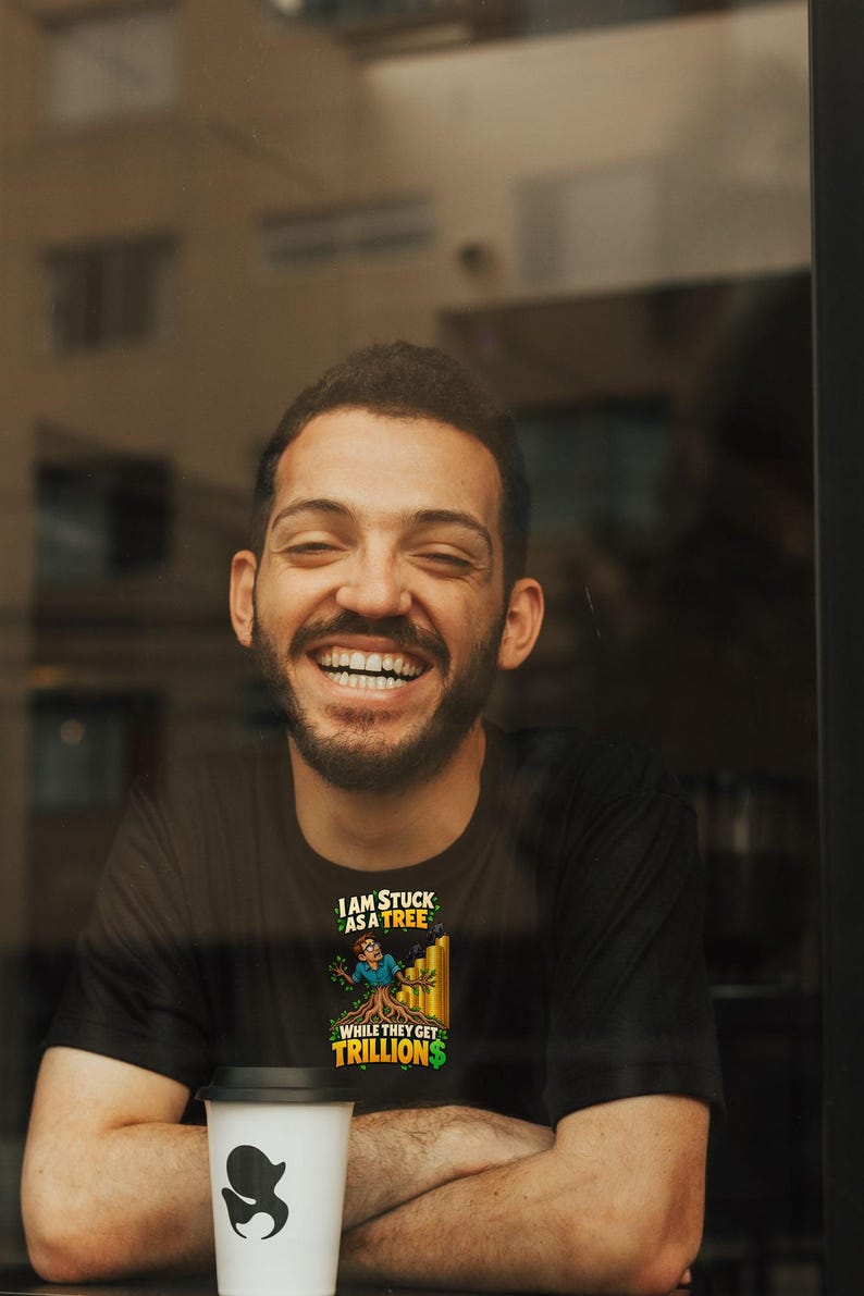 May include: A black t-shirt featuring a graphic of a person partially transformed into a tree, with the text "I AM STUCK AS A TREE WHILE THEY GET TRILLIONS" in yellow and white lettering, positioned above a white cup with a black logo.