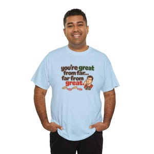 May include: A light blue short-sleeve t-shirt featuring the text "you're great from far... far from great." in brown and green, accompanied by a cartoon illustration of a man pointing to his head and small red shoe prints.