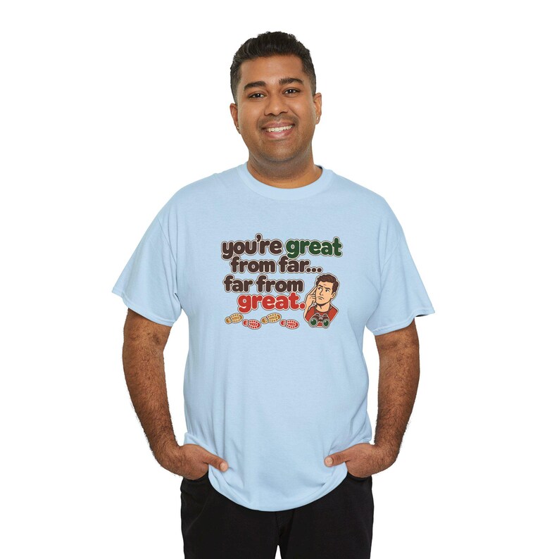 May include: A light blue short-sleeve t-shirt featuring the text "you're great from far... far from great." in brown and green, accompanied by a cartoon illustration of a man pointing to his head and small red shoe prints.