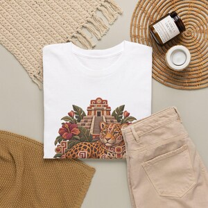 May include: A white t-shirt featuring a graphic print of a spotted jaguar resting in front of a stepped pyramid structure, surrounded by red hibiscus flowers and green tropical leaves. The design incorporates geometric patterns in earthy tones.