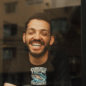 May include: A black t-shirt featuring a graphic of a fish jumping through blue waves with the white text "CURRENTLY FIGHTING THE CURRENT." The shirt is worn by a person with a beard, and a white coffee cup with a black lid is in the foreground.