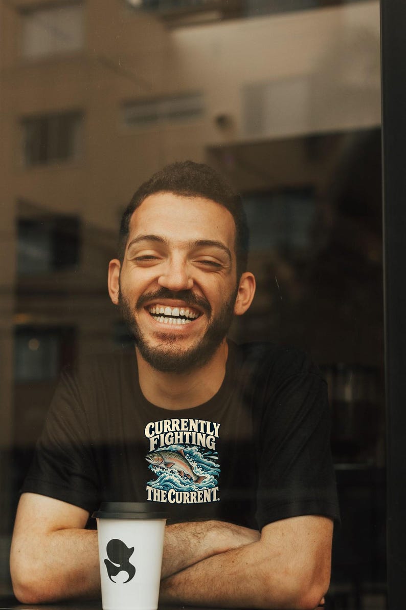 May include: A black t-shirt featuring a graphic of a fish jumping through blue waves with the white text "CURRENTLY FIGHTING THE CURRENT." The shirt is worn by a person with a beard, and a white coffee cup with a black lid is in the foreground.