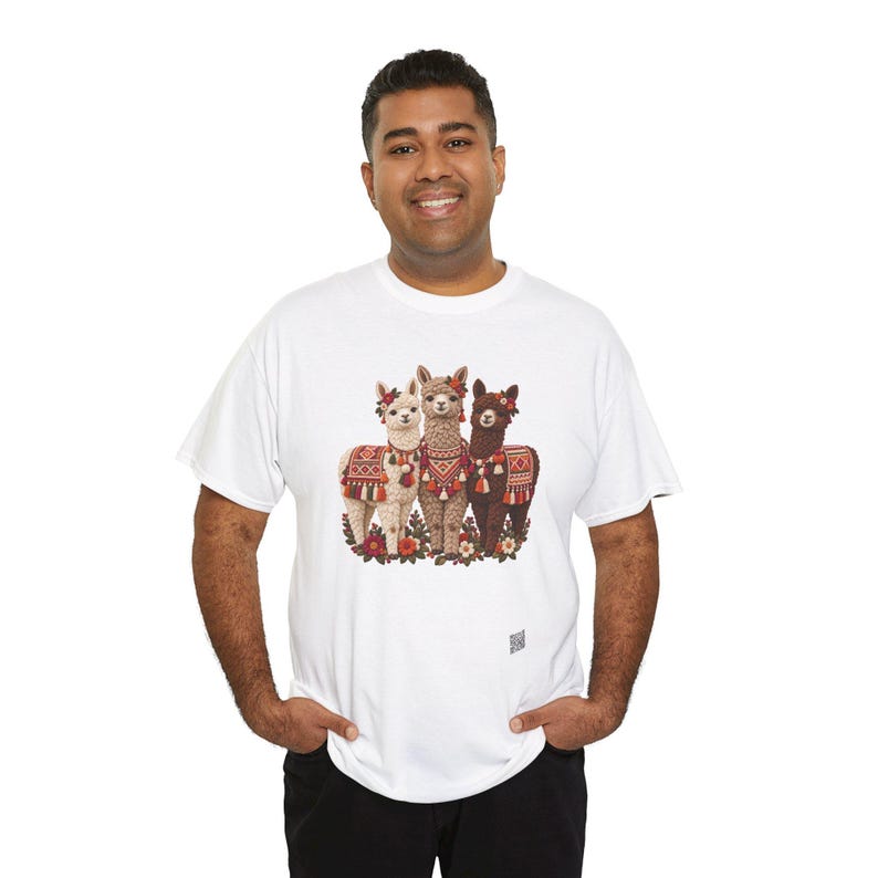 May include: A white short-sleeved t-shirt featuring a central graphic of three llamas standing together. The llamas are adorned with colorful patterned blankets, tassels, and floral headpieces in shades of red, orange, and green.