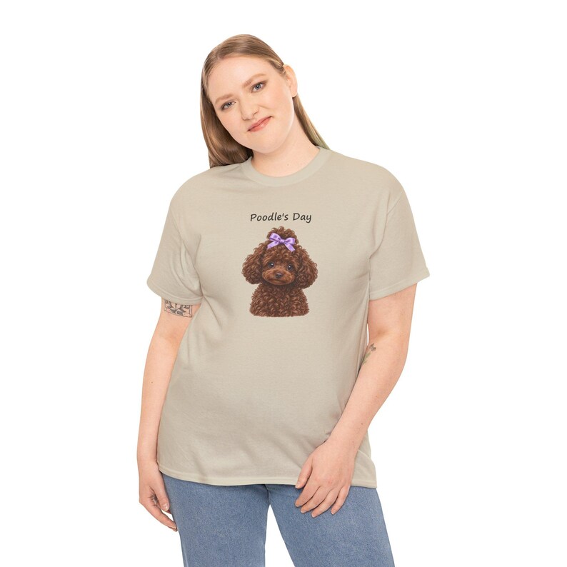May include: A beige short-sleeved t-shirt featuring a graphic of a brown curly-haired poodle with a purple bow on its head. Above the poodle, the text Poodle's Day is printed in a simple black font. The shirt has a crew neckline.