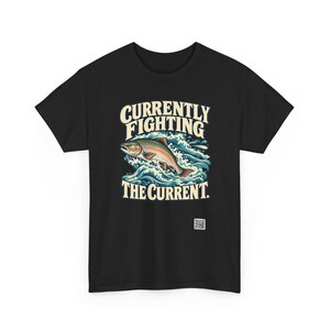 May include: A black t-shirt featuring a graphic of a trout jumping through blue waves. The text above the fish reads "CURRENTLY FIGHTING" and the text below reads "THE CURRENT." A small white square QR code is printed near the bottom right.