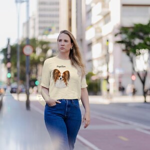 Puede incluir: Una camiseta de manga corta color crema con una ilustraci&oacute;n gr&aacute;fica de un perro Papillon marr&oacute;n y blanco con la palabra Papillon impresa sobre la cabeza del perro, combinada con unos vaqueros azules.