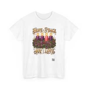 May include: A white t-shirt featuring a graphic of an Advent wreath with four lit candles in purple and pink, surrounded by evergreen branches and ribbons. The text HOPE PEACE is printed above the candles, and JOY LOVE is printed below in gold.