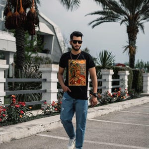 May include: A black short-sleeved t-shirt featuring a colorful graphic print of an Aztec warrior wearing an elaborate feathered headdress, paired with light blue denim jeans and white athletic sneakers with black stripes.