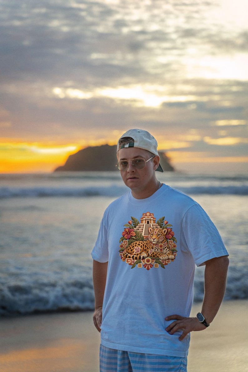 May include: A white short-sleeved t-shirt featuring a central graphic print of a resting jaguar positioned in front of a stepped pyramid, surrounded by tropical flowers and geometric patterns in shades of orange, red, and green.