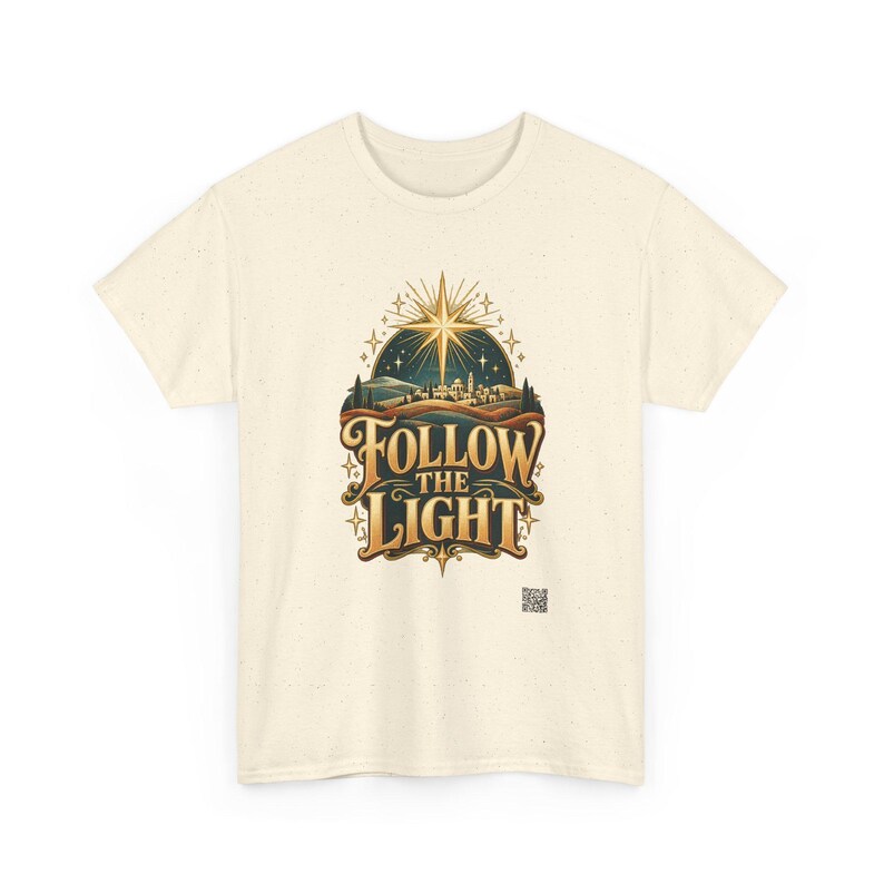 May include: A cream-colored short-sleeve t-shirt featuring a graphic print of a glowing star above a landscape with the text "FOLLOW THE LIGHT" in a stylized gold font. The design includes dark blue and brown tones with small star accents.