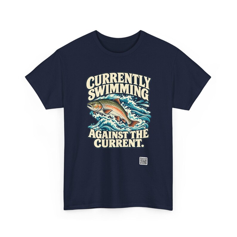 May include: A navy blue t-shirt featuring a graphic of a trout jumping through blue and white waves. The text above the fish reads CURRENTLY SWIMMING and the text below reads AGAINST THE CURRENT. A small white QR code is printed near the bottom.