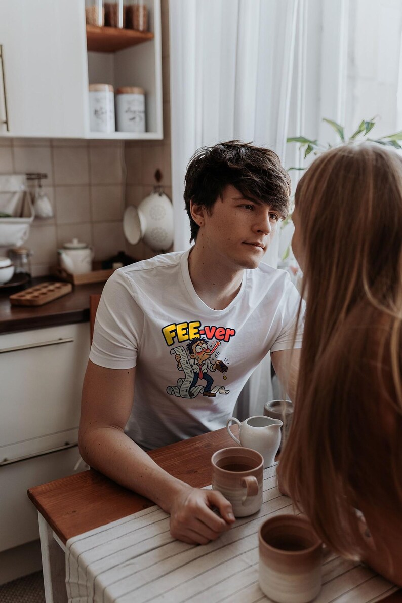 May include: A white short-sleeved t-shirt featuring a graphic print of a cartoon character with a thermometer in their mouth, holding a long receipt, with the text FEE-ver printed in yellow and red letters above the character.
