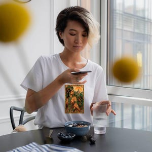 May include: A white short-sleeved t-shirt featuring a rectangular graphic print on the chest. The illustration depicts a person wearing ornate traditional attire and a headdress, set against a backdrop of a pyramid and lush green foliage.