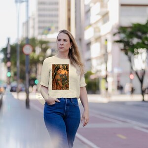 May include: A cream-colored short-sleeve t-shirt featuring a rectangular graphic print of a person wearing traditional attire and jewelry, set against a warm-toned background with foliage and architectural elements.