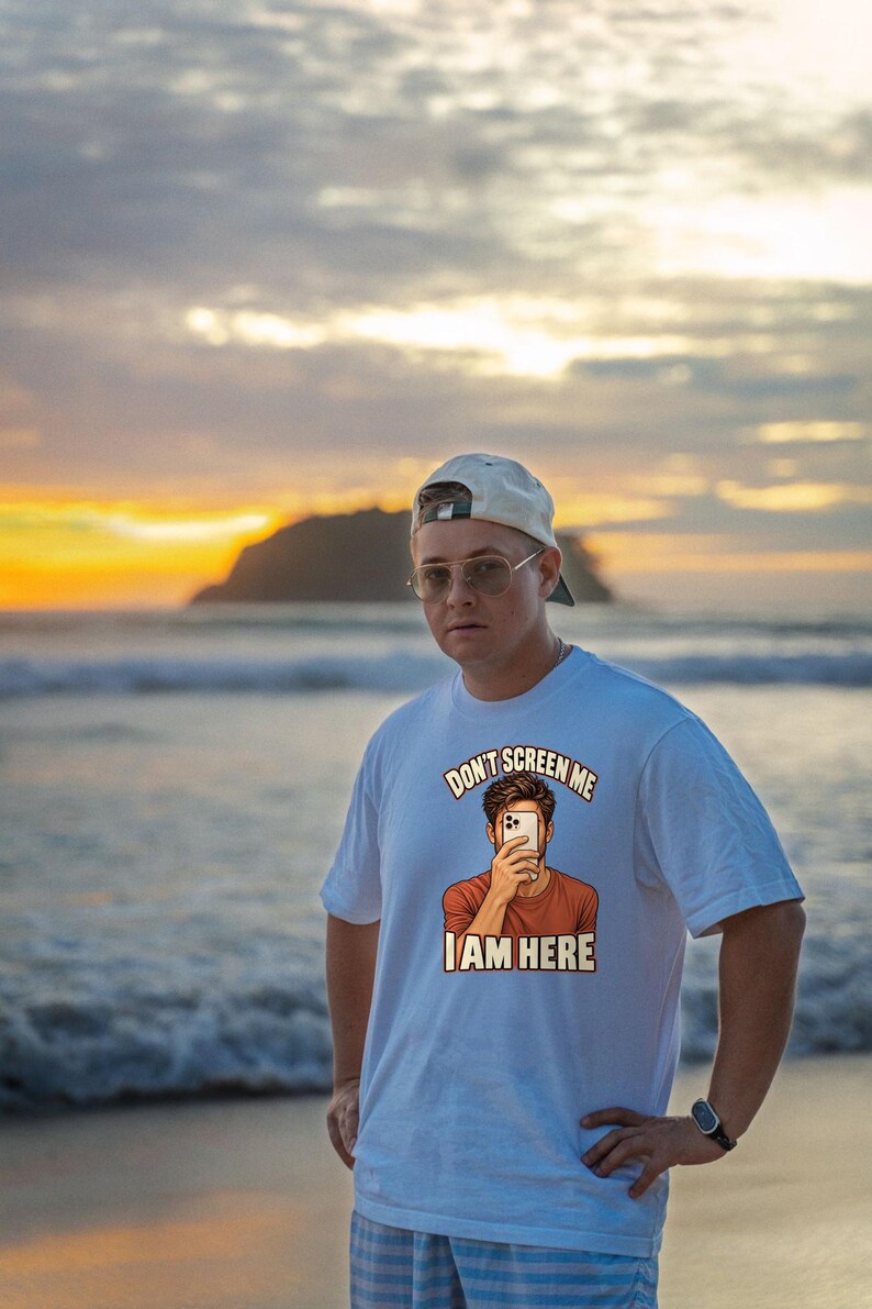Op de afbeelding: Een wit T-shirt met korte mouwen en een grafische illustratie van een persoon die een smartphone voor het gezicht houdt. De tekst boven de illustratie luidt DON'T SCREEN ME en daaronder staat I AM HERE in vetgedrukte letters.