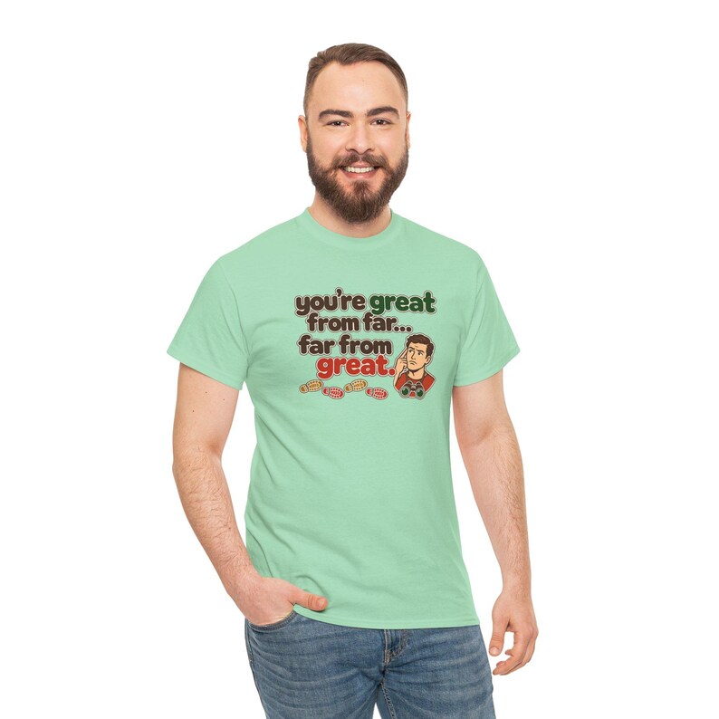 May include: A light green short-sleeve t-shirt featuring the text "you're great from far... far from great." in dark brown and red lettering, accompanied by a cartoon illustration of a person and small red gaming controllers.