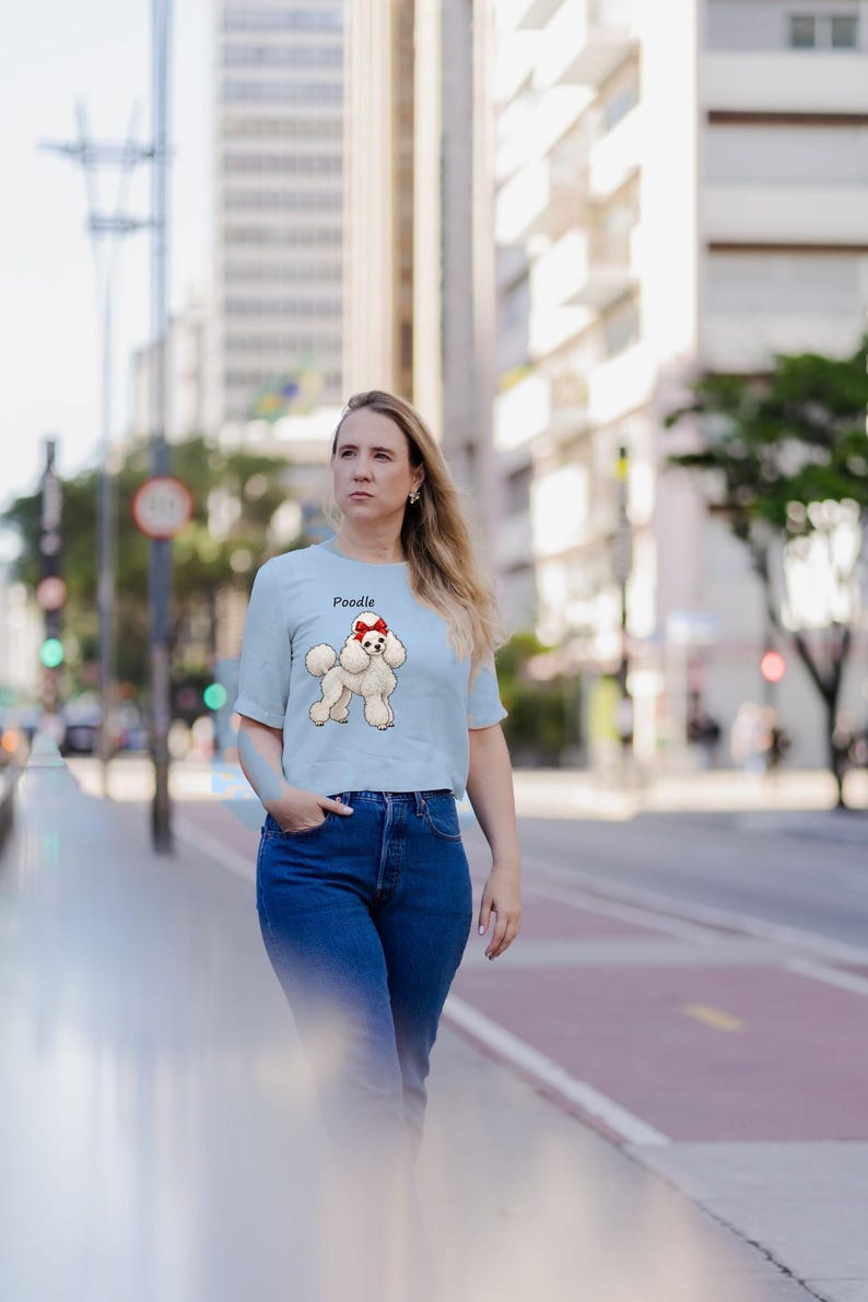 May include: A light blue short-sleeved t-shirt featuring a graphic illustration of a white poodle with a red bow on its head. The word Poodle is printed in black text above the dog graphic. The shirt has a crew neckline and a straight hem.