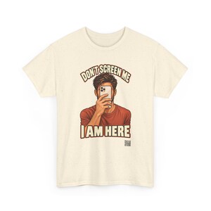 Op de afbeelding: Een cr&egrave;mekleurig T-shirt met korte mouwen en een afbeelding van een persoon die een smartphone voor het gezicht houdt. De tekst erboven luidt "DON'T SCREEN ME" en eronder "I AM HERE" in vetgedrukte letters. Een kleine QR-code is rechtsonder zichtbaar.