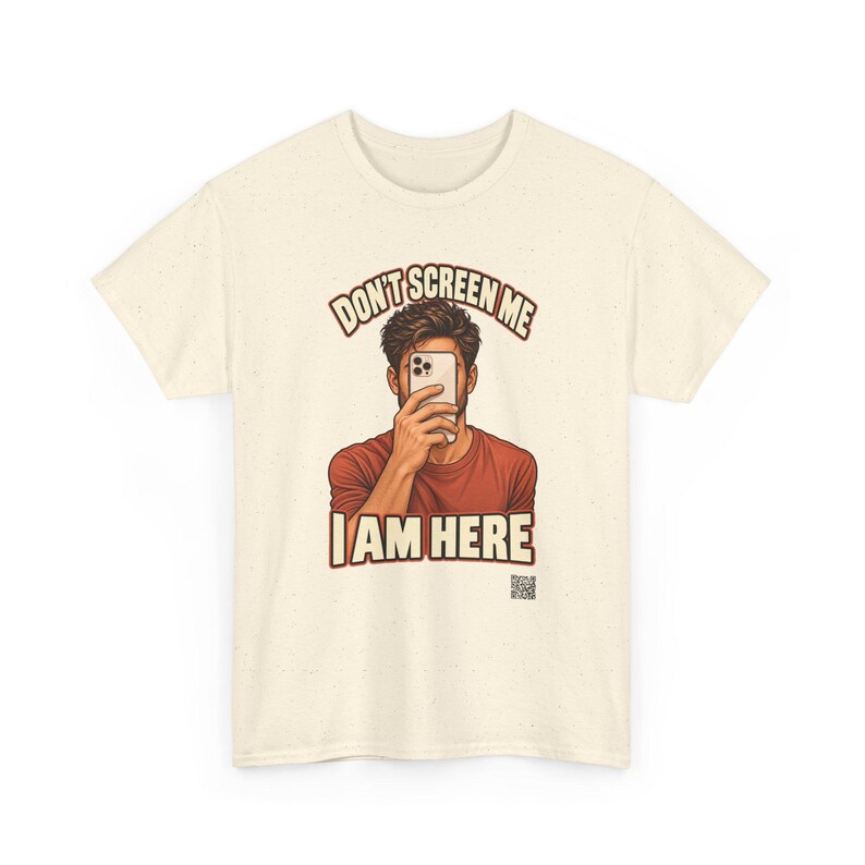 Op de afbeelding: Een cr&egrave;mekleurig T-shirt met korte mouwen en een afbeelding van een persoon die een smartphone voor het gezicht houdt. De tekst erboven luidt "DON'T SCREEN ME" en eronder "I AM HERE" in vetgedrukte letters. Een kleine QR-code is rechtsonder zichtbaar.