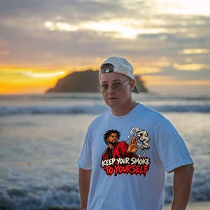 May include: A white short-sleeved t-shirt featuring a graphic illustration of a person in a red jacket holding a hand out with a smoking cigarette. The text "KEEP YOUR SMOKE TO YOURSELF" is printed in bold, stylized red and black lettering below the figure.