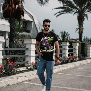 May include: A black short-sleeved t-shirt featuring a graphic of a purple and green robot with the text "AUTOPILOT MODE" and "CLEAR THE WAY!" paired with blue denim jeans and white sneakers, worn with dark sunglasses and a gold chain.