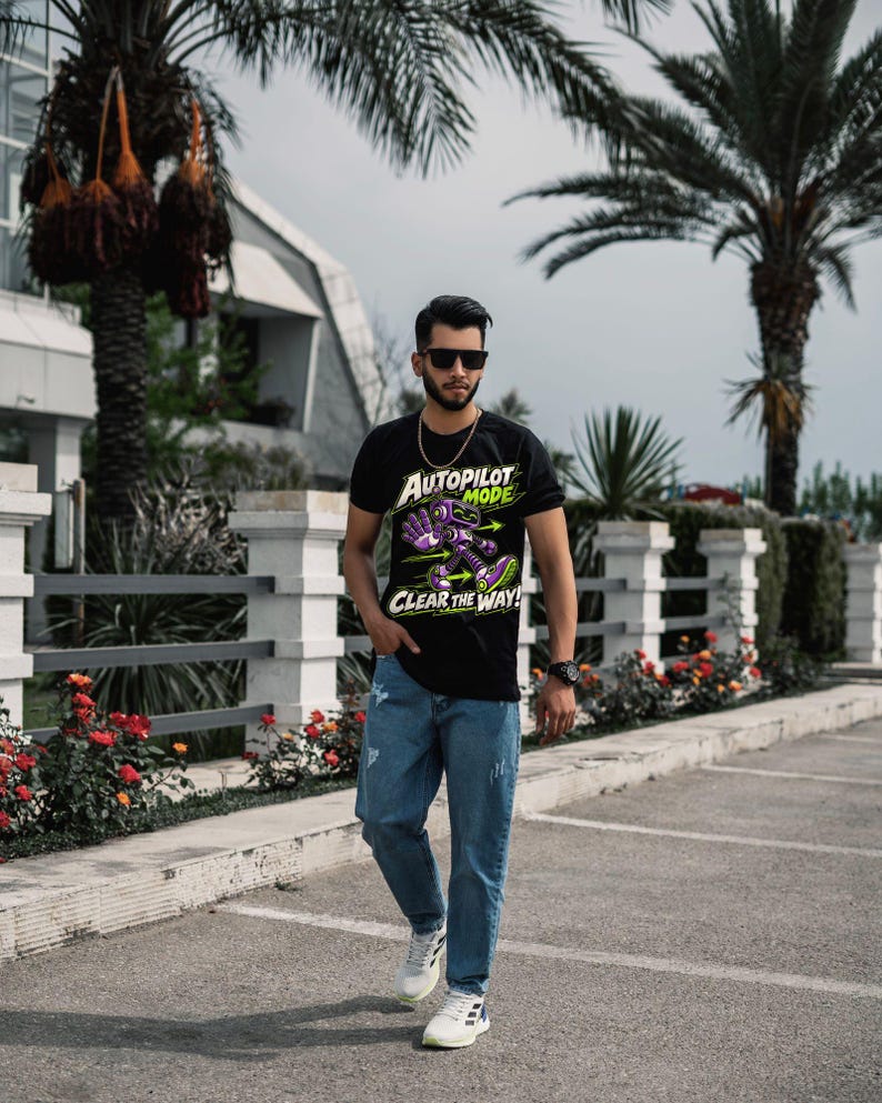 May include: A black short-sleeved t-shirt featuring a graphic of a purple and green robot with the text "AUTOPILOT MODE" and "CLEAR THE WAY!" paired with blue denim jeans and white sneakers, worn with dark sunglasses and a gold chain.