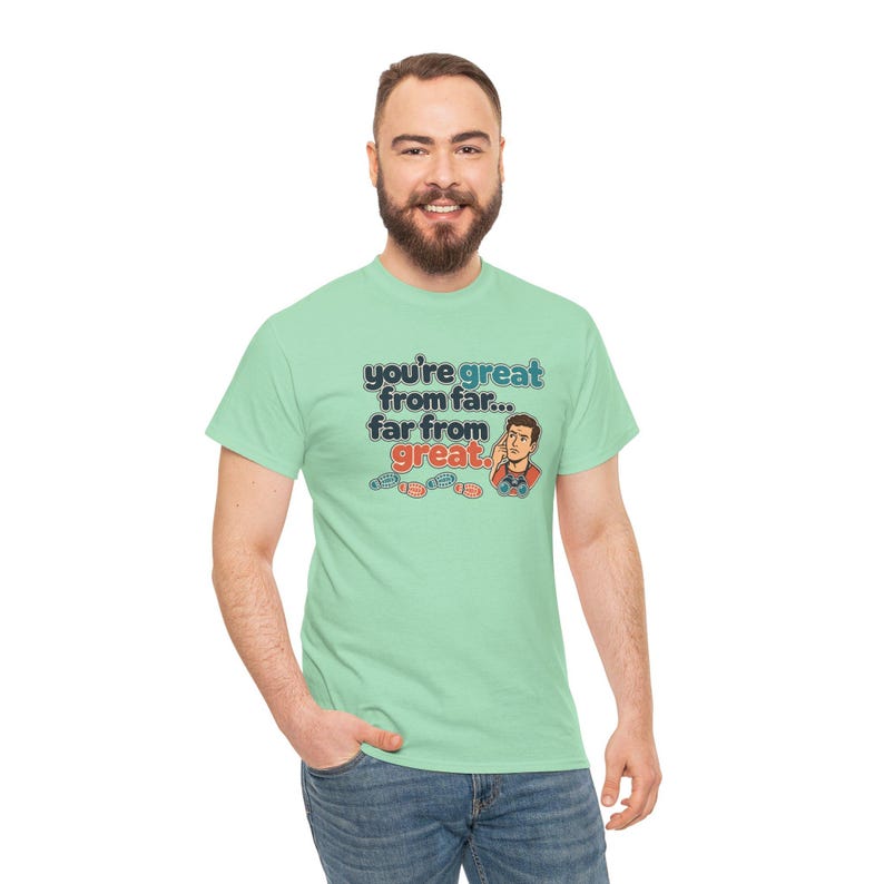 May include: A light green t-shirt featuring the text "you're great from far... far from great." in blue and red lettering, accompanied by a cartoon illustration of a person looking through binoculars and small footprint icons.