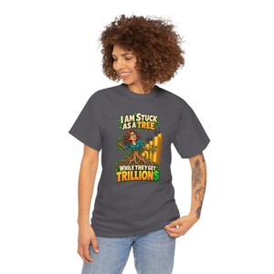 May include: A dark gray t-shirt featuring a graphic of a person with brown hair merging into a tree trunk. The text reads I AM STUCK AS A TREE WHILE THEY GET TRILLIONS, accompanied by an illustration of rising gold bars in the background.