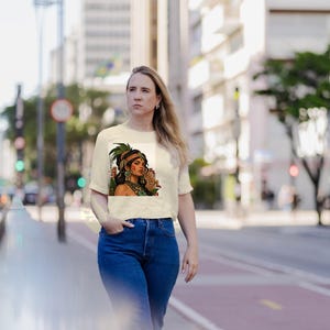 Puede incluir: Una camiseta de manga corta color crema con un estampado gr&aacute;fico de una persona de cabello oscuro, que lleva un tocado de plumas verdes, joyas doradas y una prenda verde sobre un fondo circular con patrones.