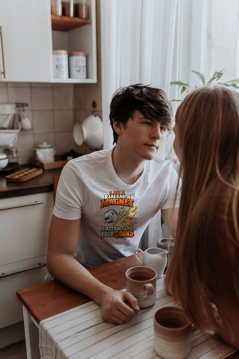 May include: A white short-sleeved t-shirt featuring a graphic of a smiling magnet with the text I AM A RARE EARTH MAGNET I ATTRACT YOUR COINS. The design includes a grey cylindrical magnet surrounded by several gold-colored coins.