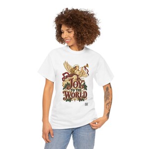 May include: A white short-sleeve t-shirt featuring a graphic of an angel playing a trumpet with red ribbons and green holly leaves. The text reads JOY TO THE WORLD in a decorative font with small star accents and a small QR code at the bottom.