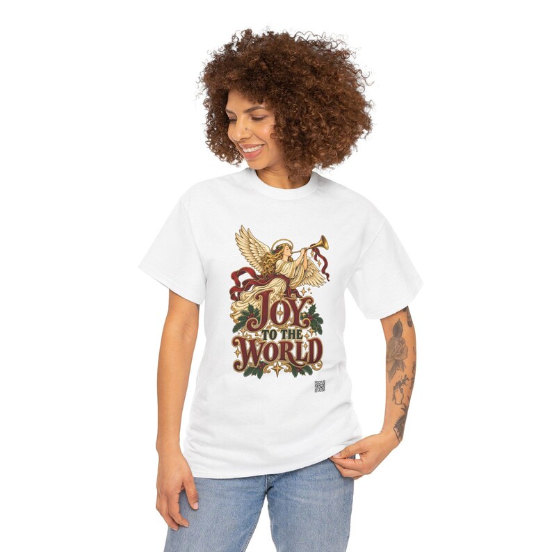 May include: A white short-sleeve t-shirt featuring a graphic of an angel playing a trumpet with red ribbons and green holly leaves. The text reads JOY TO THE WORLD in a decorative font with small star accents and a small QR code at the bottom.