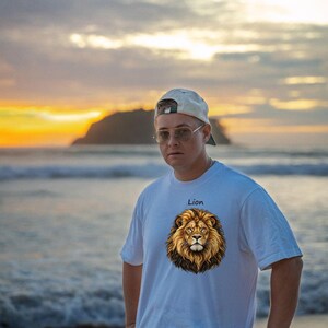 May include: A white short-sleeved t-shirt featuring a graphic print of a lion's head in the center of the chest with the word Lion printed in black text above the lion illustration. The shirt has a crew neckline and a standard fit.