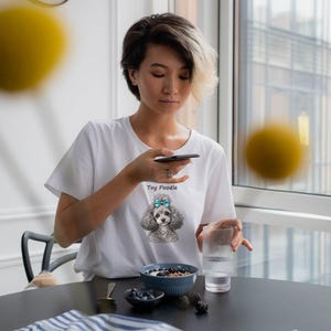 K&ouml;nnte beinhalten: Ein wei&szlig;es Kurzarm-T-Shirt mit einer grafischen Illustration eines grauen Toypudels mit einer leuchtend blauen Schleife auf dem Kopf, platziert &uuml;ber dem Text Toy Poodle, der in einer einfachen schwarzen Schrift auf der Brust gedruckt ist.