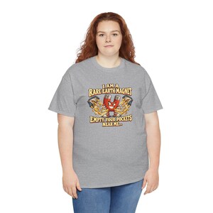 Op de afbeelding: Een grijs gem&ecirc;leerd t-shirt met een cartoonfiguur van een rode hoefijzermagneet die gouden munten uit twee zakken trekt. De tekst luidt: I AM A RARE EARTH MAGNET EMPTY YOUR POCKETS NEAR ME...