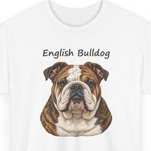 Puede incluir: Una camiseta blanca con una ilustraci&oacute;n gr&aacute;fica de la cabeza y los hombros de un bulldog ingl&eacute;s. El perro tiene pelaje marr&oacute;n y blanco, cara arrugada y ojos oscuros. El texto "English Bulldog" est&aacute; impreso en negro sobre la cabeza.