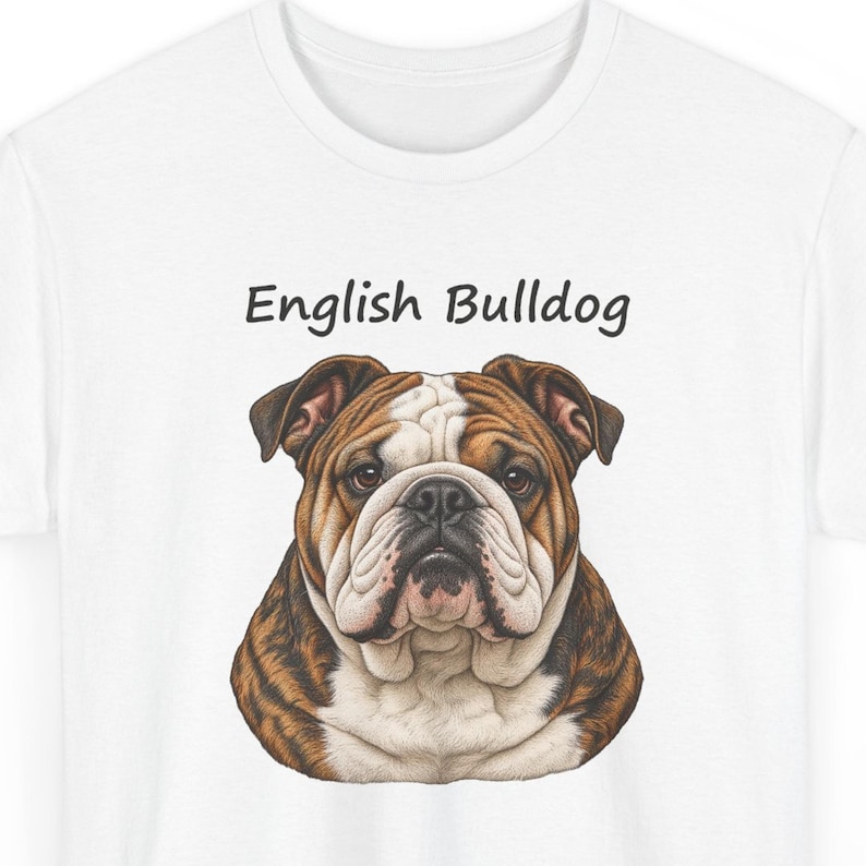 Puede incluir: Una camiseta blanca con una ilustraci&oacute;n gr&aacute;fica de la cabeza y los hombros de un bulldog ingl&eacute;s. El perro tiene pelaje marr&oacute;n y blanco, cara arrugada y ojos oscuros. El texto "English Bulldog" est&aacute; impreso en negro sobre la cabeza.