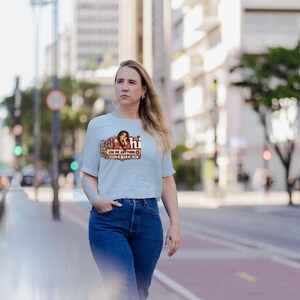 May include: A light blue short-sleeved t-shirt featuring a graphic print of a person with long dark hair and the text "hi" and "ASK ME ANYTHING" in a decorative frame. The shirt is paired with dark blue denim jeans with a classic fit.