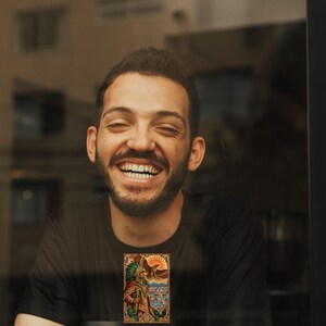 Pu&ograve; includere: Una t-shirt nera a maniche corte con una stampa grafica rettangolare raffigurante un guerriero azteco con copricapo di piume, un'aquila e un sole sopra un paesaggio urbano. &Egrave; presente un bicchiere da caff&egrave; monouso bianco con coperchio nero.