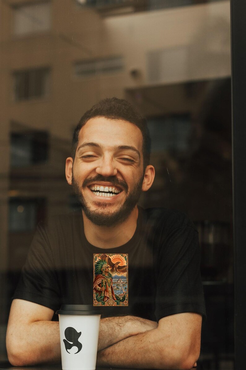 Pu&ograve; includere: Una t-shirt nera a maniche corte con una stampa grafica rettangolare raffigurante un guerriero azteco con copricapo di piume, un'aquila e un sole sopra un paesaggio urbano. &Egrave; presente un bicchiere da caff&egrave; monouso bianco con coperchio nero.