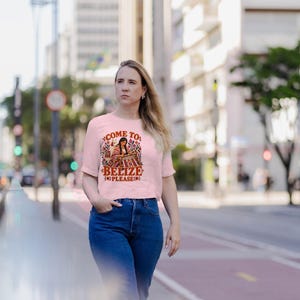 May include: A light pink short-sleeved t-shirt featuring a graphic illustration of a person weaving, surrounded by colorful floral patterns and the text "COME TO BELIZE PLEASE" in a bold, dark red font on the front of the garment.