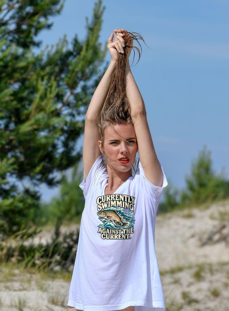 May include: A white short-sleeved t-shirt featuring a graphic of a fish jumping out of blue water waves with the text "CURRENTLY SWIMMING AGAINST THE CURRENT" printed in a bold, dark font above and below the illustration.