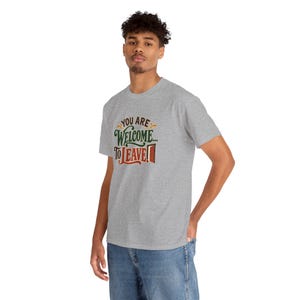 May include: A heather gray short-sleeve t-shirt featuring the text "YOU ARE WELCOME... TO LEAVE!" in a mix of brown, green, and red lettering, with a small red door icon next to the word LEAVE, paired with blue denim jeans.
